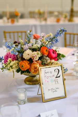 Colorful wedding centerpiece with ranunculus and delphinium beside a gold-framed table number sign reading love is forever 2.