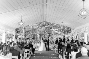 Outdoor wedding ceremony under a white pavilion surrounded by lush green trees with guests seated in white chairs.