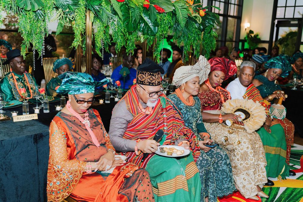Guests in vibrant traditional African attire celebrate at a colorful cultural ceremony with lush green decorations.