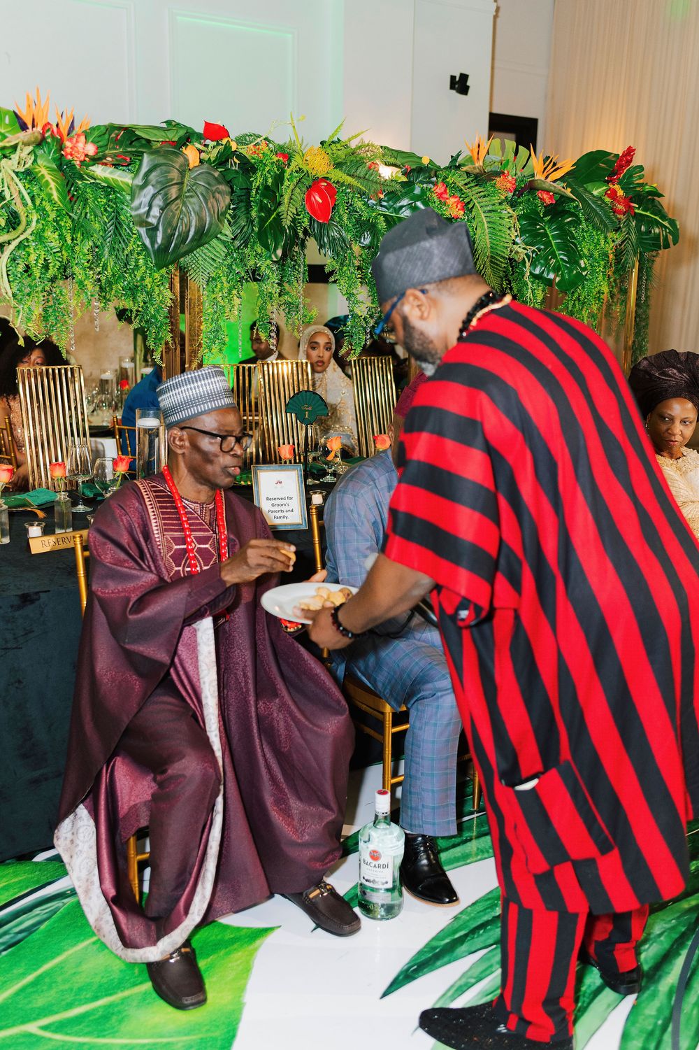 Two men in vibrant traditional Nigerian attire share a moment at a festive cultural celebration with tropical floral decor.