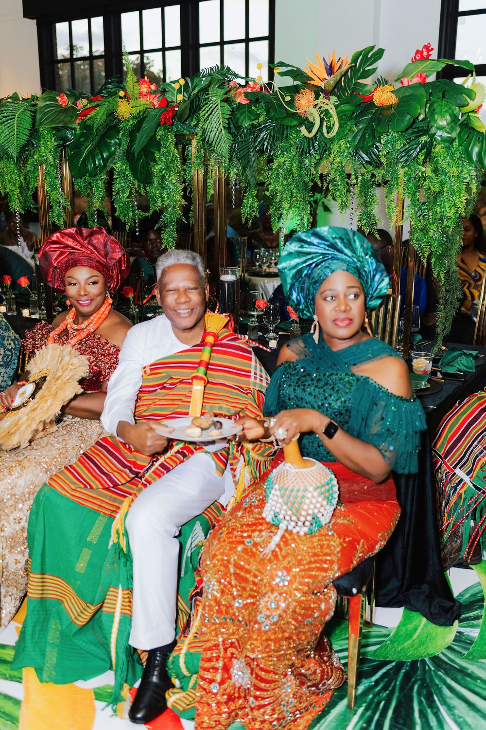 Three people in vibrant African traditional attire seated at a colorful cultural celebration with tropical floral decor.