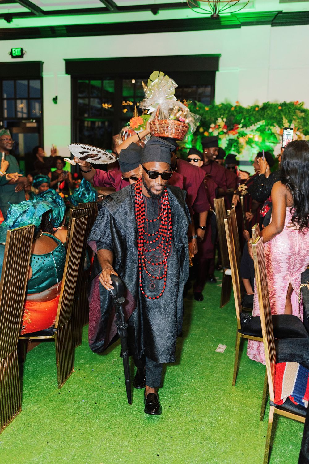 Man in traditional African attire with beaded necklaces and sunglasses walks down aisle at cultural wedding ceremony.