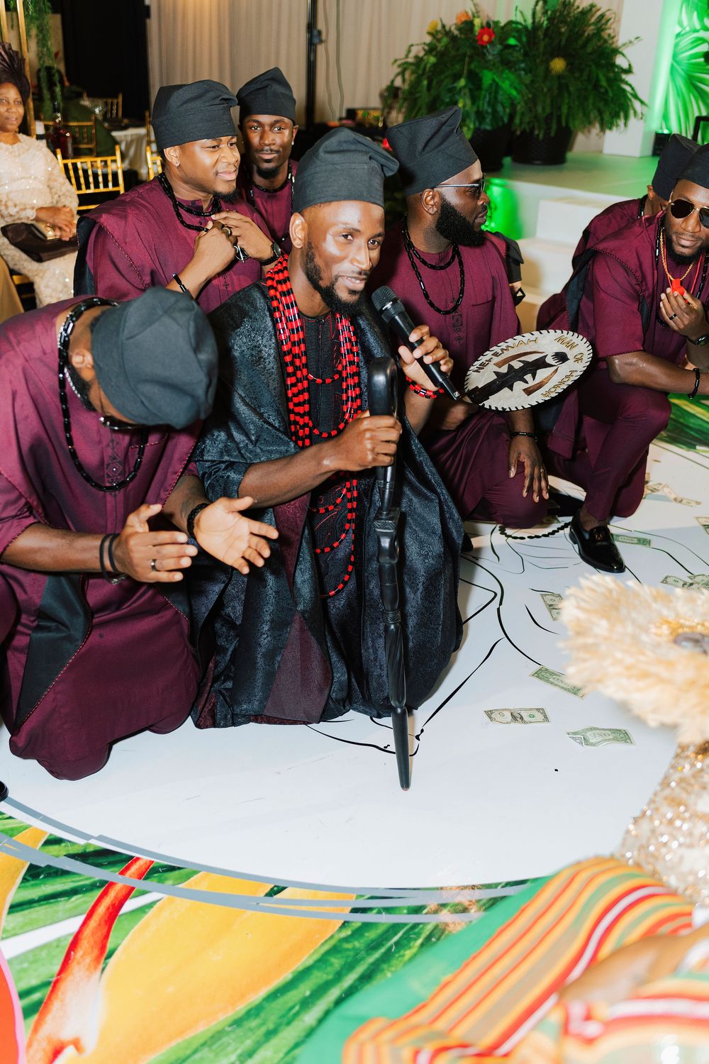 Men in traditional African attire perform at a cultural celebration, one singing into a microphone on a decorated stage.