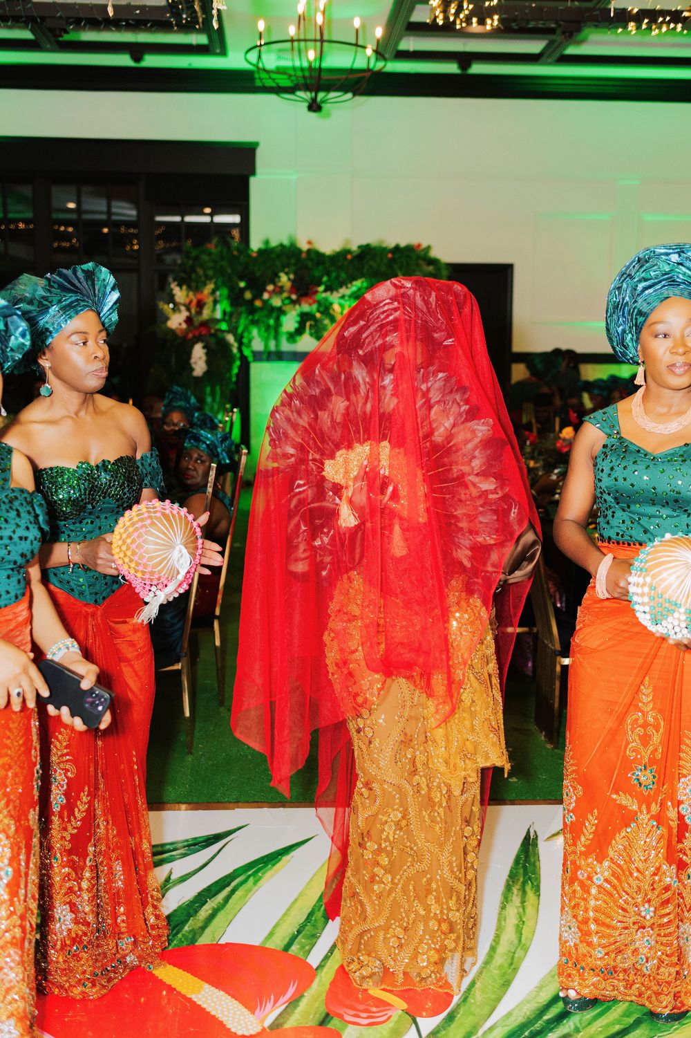 Nigerian traditional wedding ceremony with bride in red veil and orange attire, flanked by bridesmaids in green and orange.