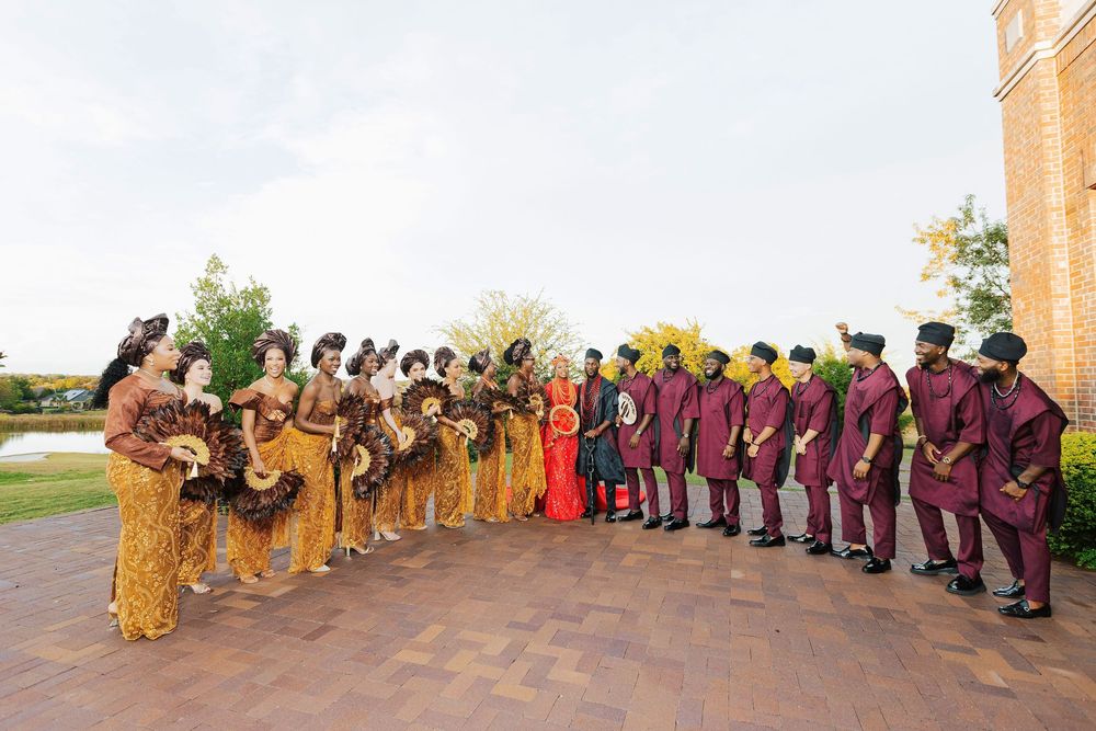 Large wedding party in vibrant African attire, women in yellow and men in burgundy, posing outdoors at elegant venue.