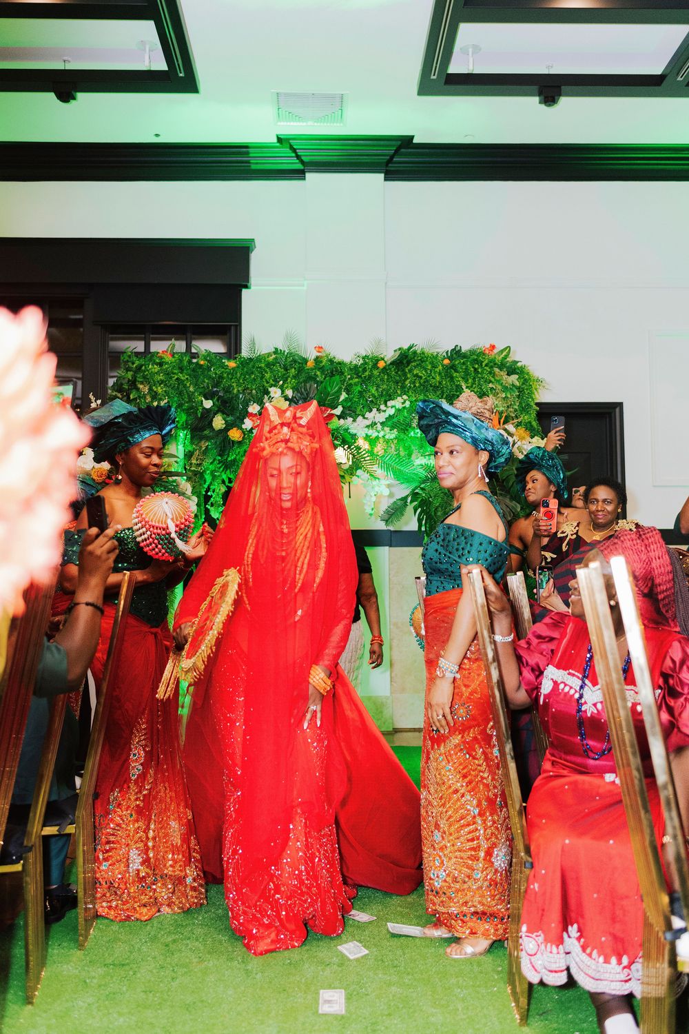 Nigerian bride in red traditional attire with veil walks down aisle surrounded by women in orange ceremonial dress.