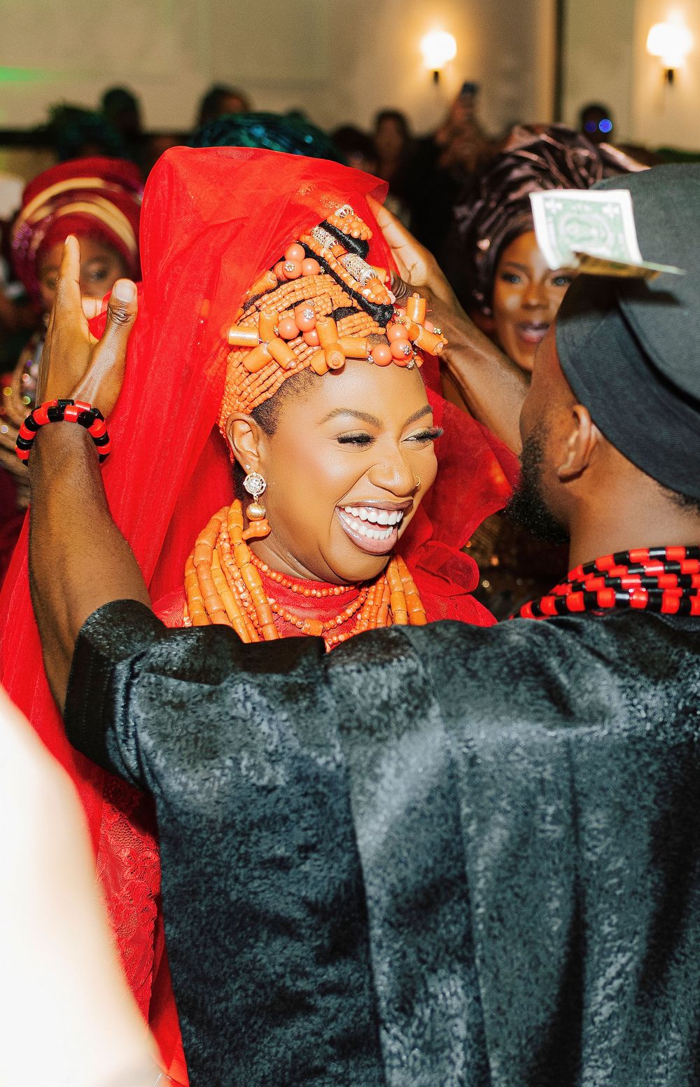 Bride in traditional Nigerian coral beaded headdress and red veil smiles joyfully during cultural wedding ceremony.
