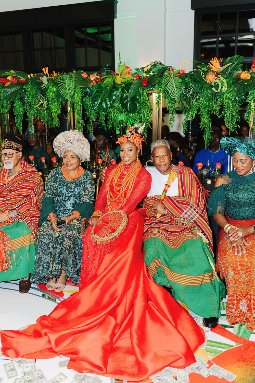 Bride in red traditional Nigerian attire with coral beads seated among women in colorful cultural dress at a traditional wedding ceremony.