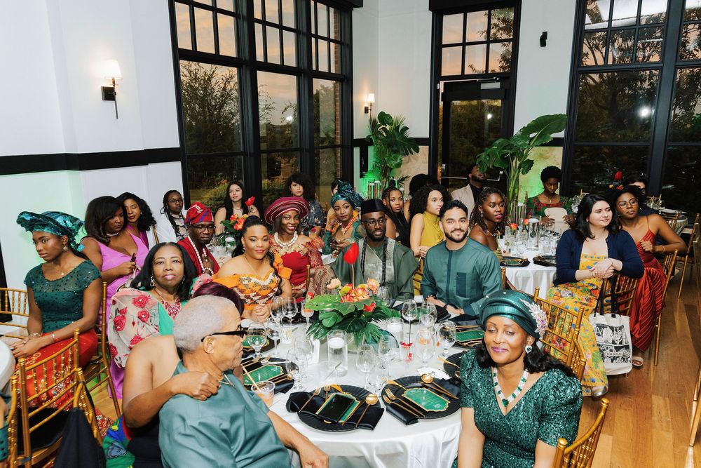 Guests in colorful African attire celebrate at a traditional wedding reception dinner with lush green decor.