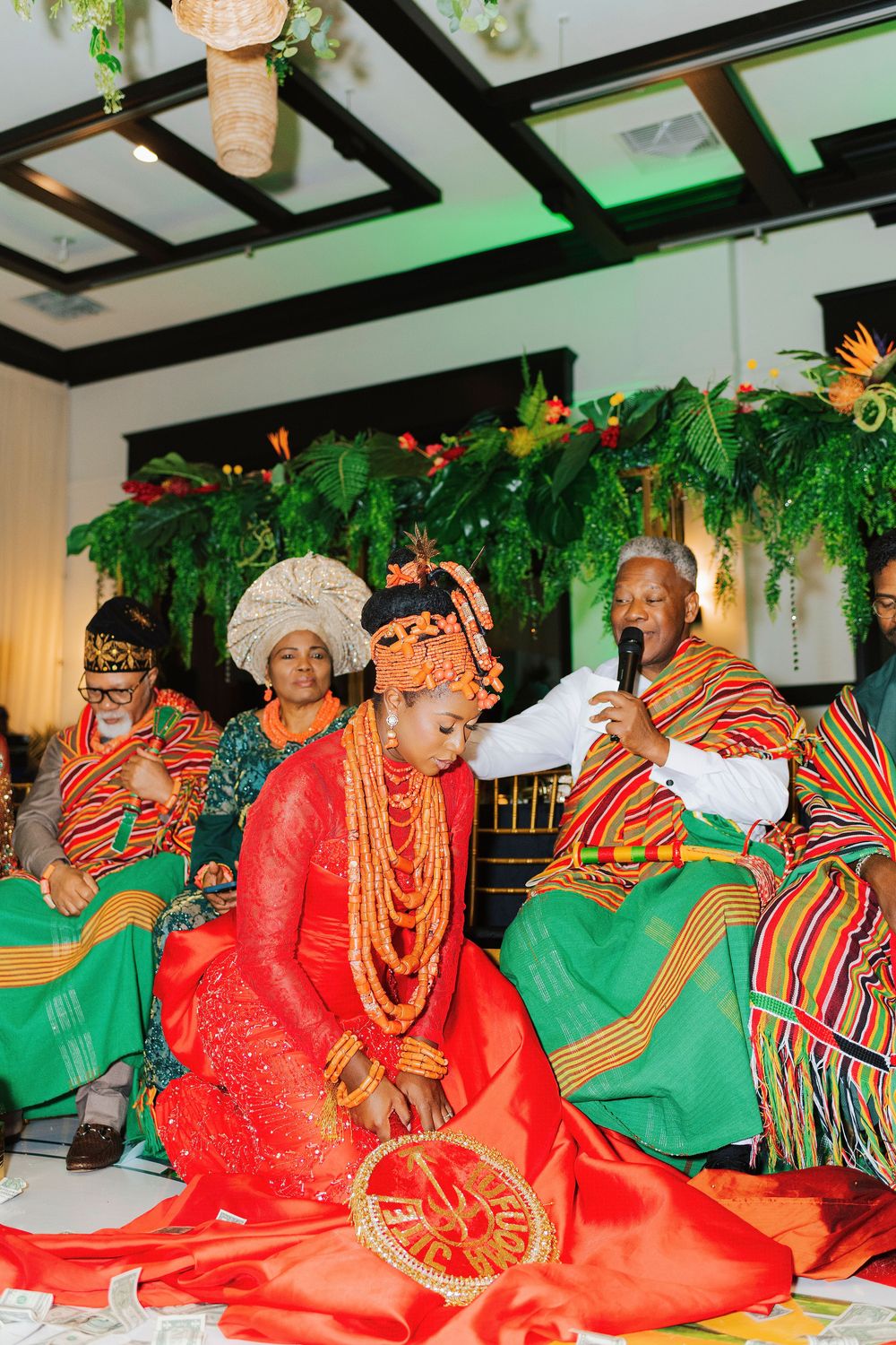 Nigerian bride in red lace gown with coral beads kneels during traditional wedding ceremony with colorful kente-clad guests.