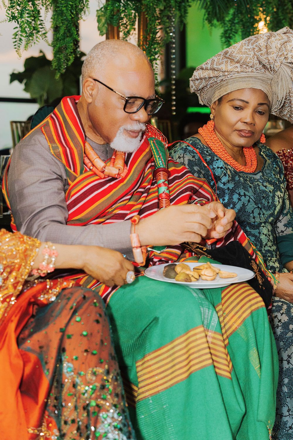 Couple in vibrant Nigerian traditional attire sharing food at a cultural ceremony, wearing coral beads and kente cloth.