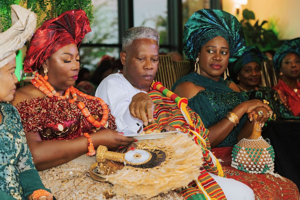 Three women in vibrant Nigerian traditional attire and gele headwraps participate in a colorful cultural ceremony.