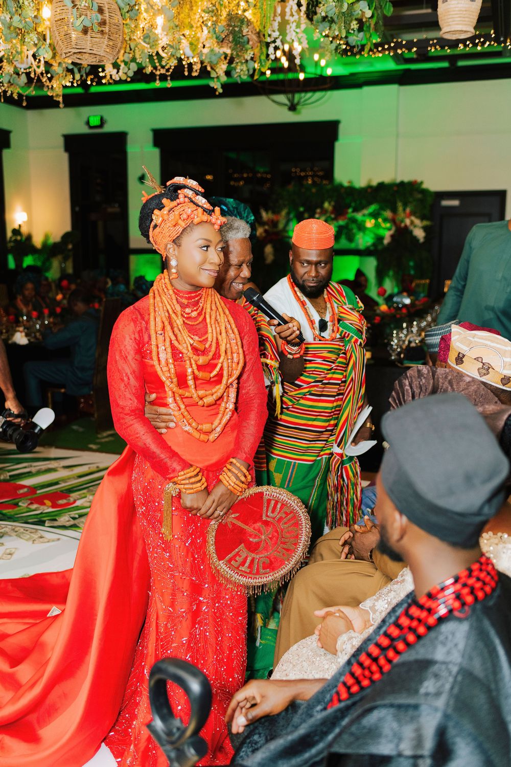 Couple in vibrant red and gold traditional Nigerian wedding attire surrounded by guests at a colorful ceremony.