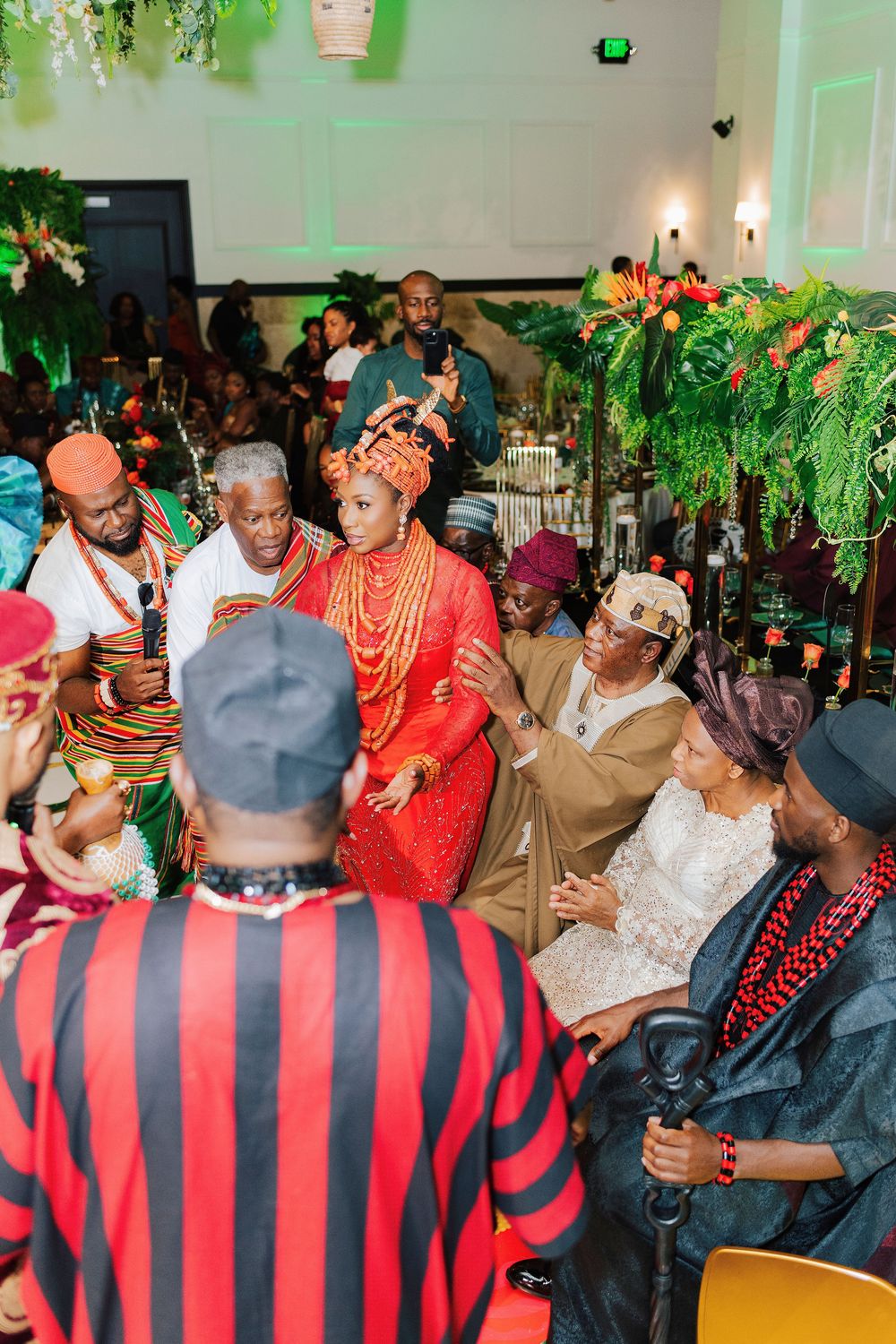 Traditional African wedding ceremony with bride in red attire and orange beaded jewelry surrounded by guests in colorful cultural dress.