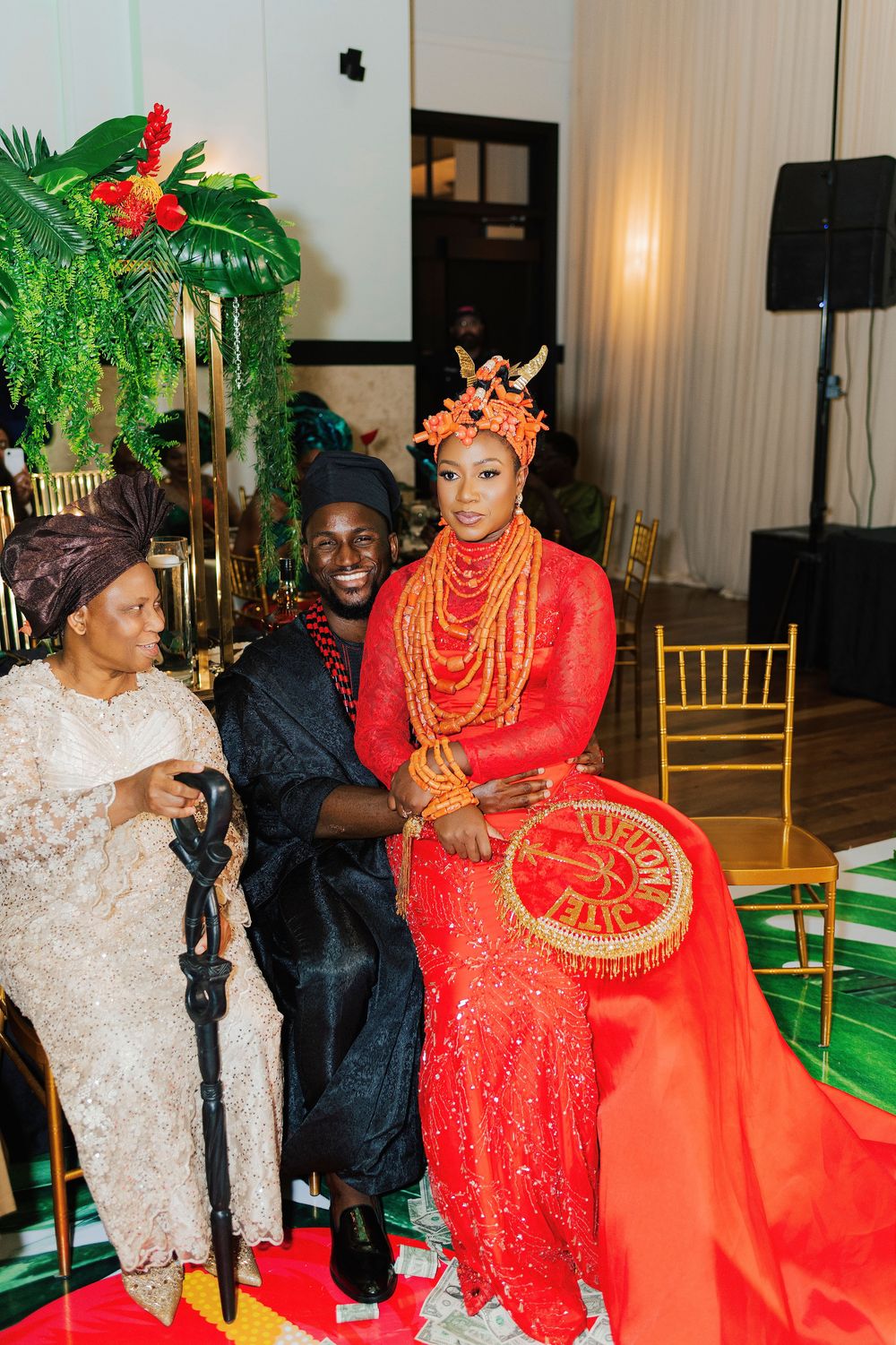 Nigerian bride in red traditional attire with coral beads and crown sits with groom in black at cultural wedding ceremony.
