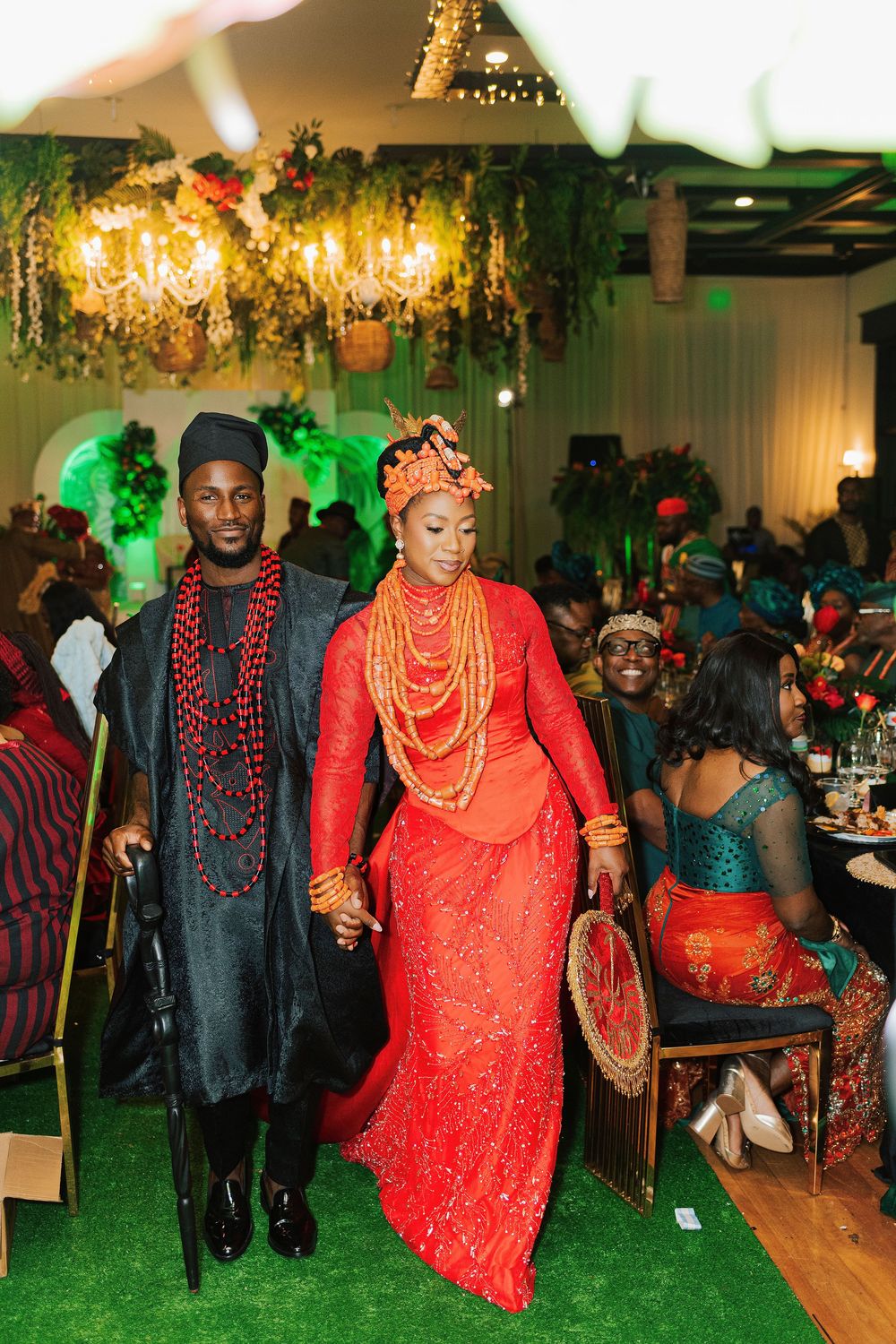 Couple in traditional Nigerian wedding attire, bride in red lace gown with coral beads, groom in dark agbada, walking at reception.