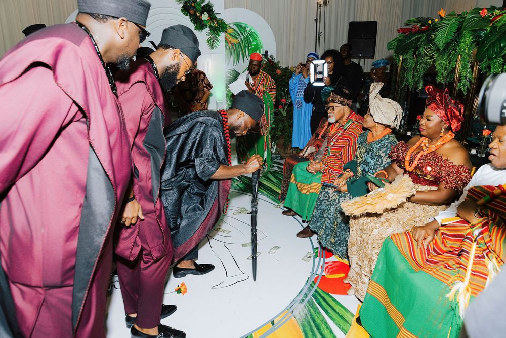 Guests in colorful traditional African attire greet seated elders at a festive cultural ceremony with tropical decor.