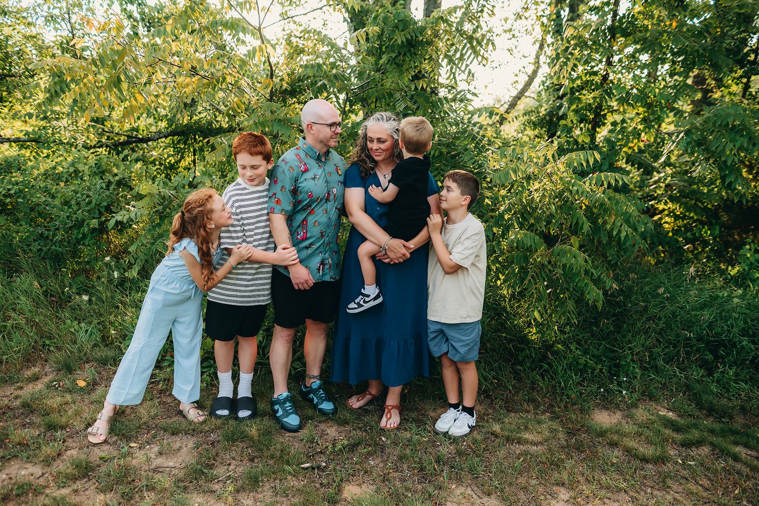 Family of five poses outdoors among lush green trees, smiling and embracing in a warm, candid portrait.
