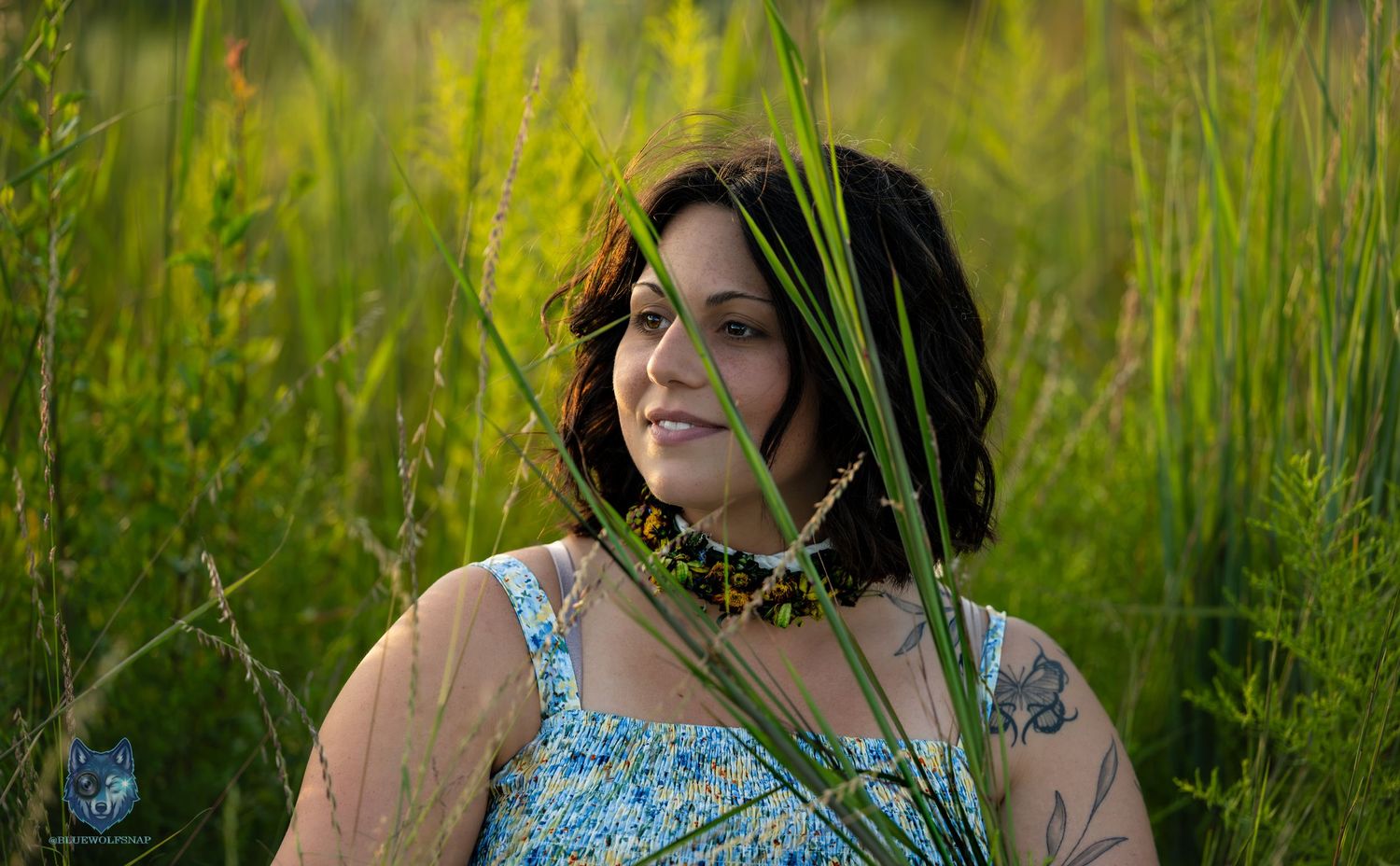 Woman in blue floral dress smiles among tall green grass in golden sunlight. Chi.Shooters meet gabbsbosh