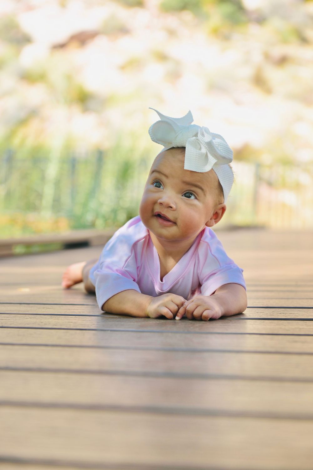 Adorable baby girl wearing a white bow headband doing tummy time on a wooden deck outdoors.