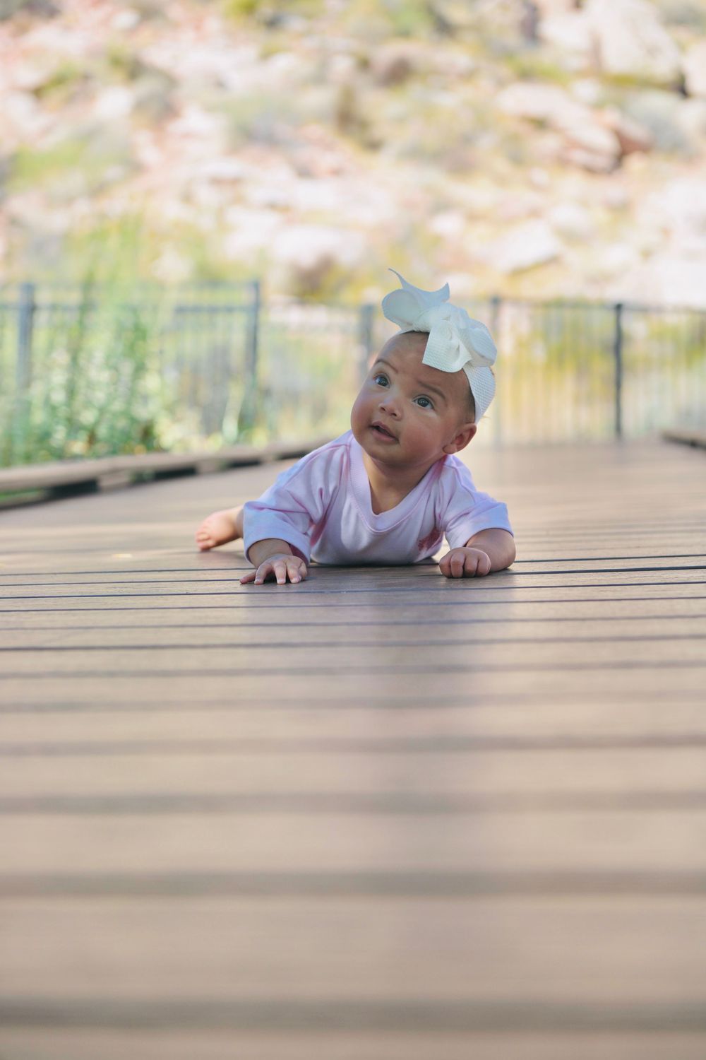 Adorable baby girl in pink onesie and bow headband lying on wooden deck outdoors, looking up curiously.