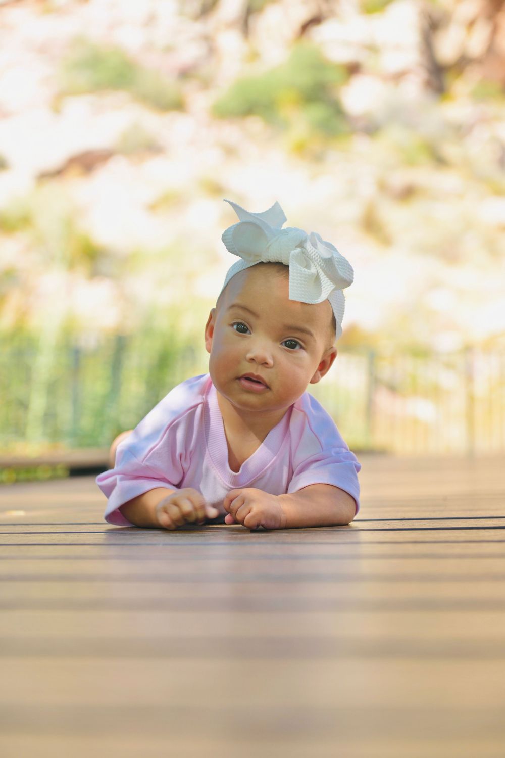 Adorable baby girl in pink outfit with white bow headband lying on wooden deck outdoors.