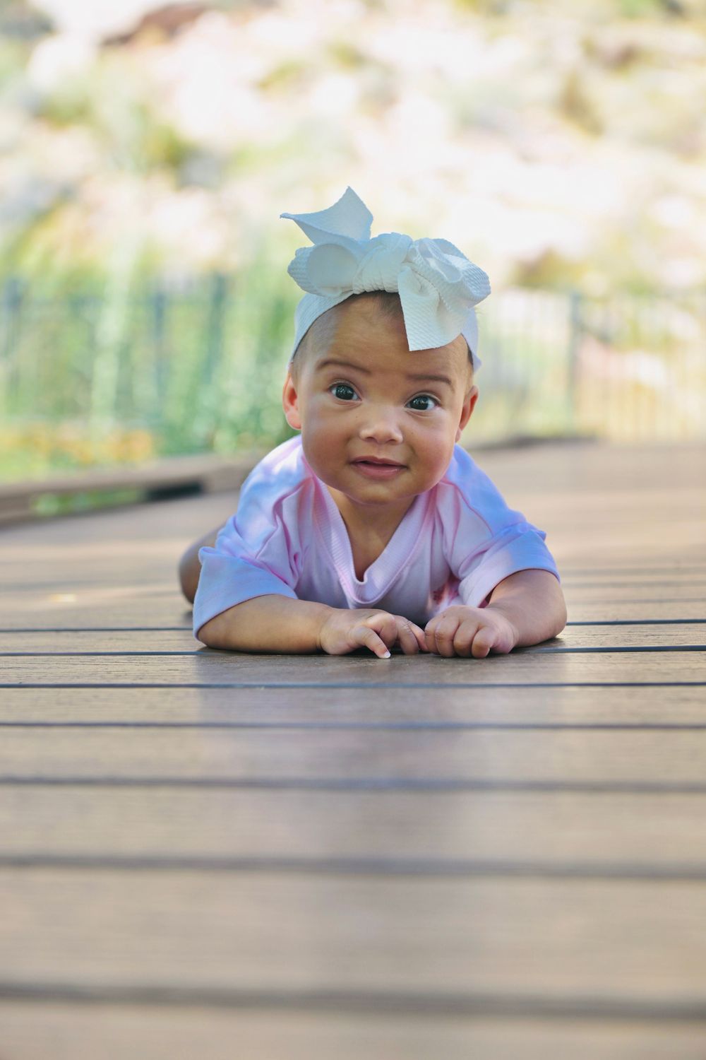 Adorable baby girl wearing a large white bow headband, lying on a wooden deck outdoors, smiling at the camera.