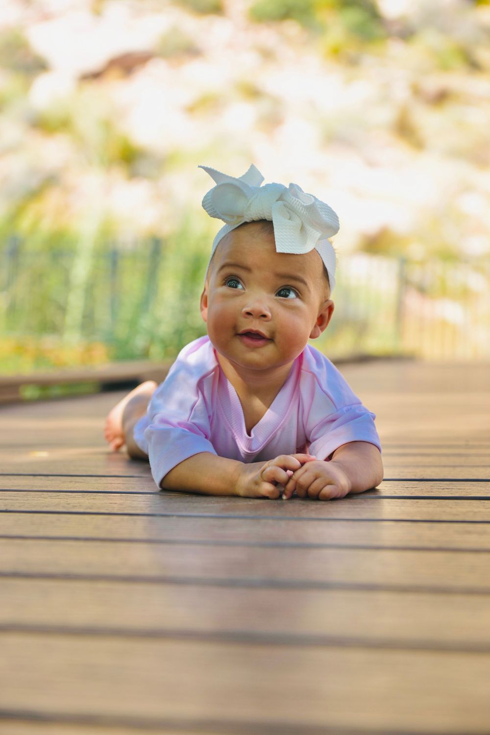 Adorable baby girl in pink outfit with white bow headband doing tummy time on wooden deck outdoors.