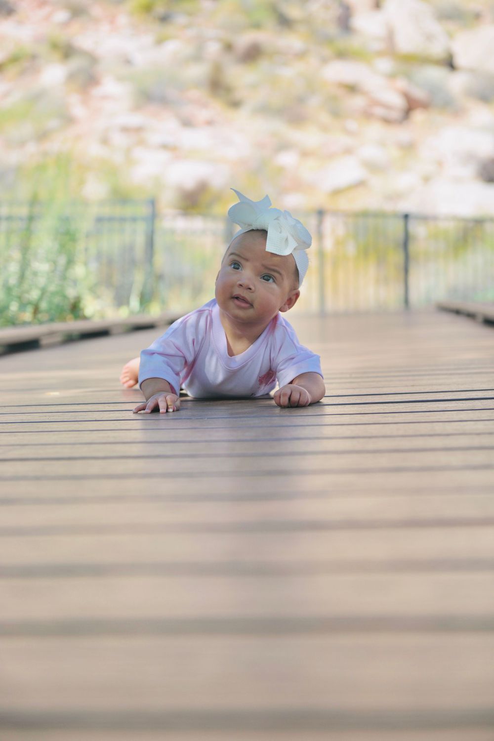 A baby girl in a pink outfit and white bow headband lies on a wooden deck, looking up curiously outdoors.