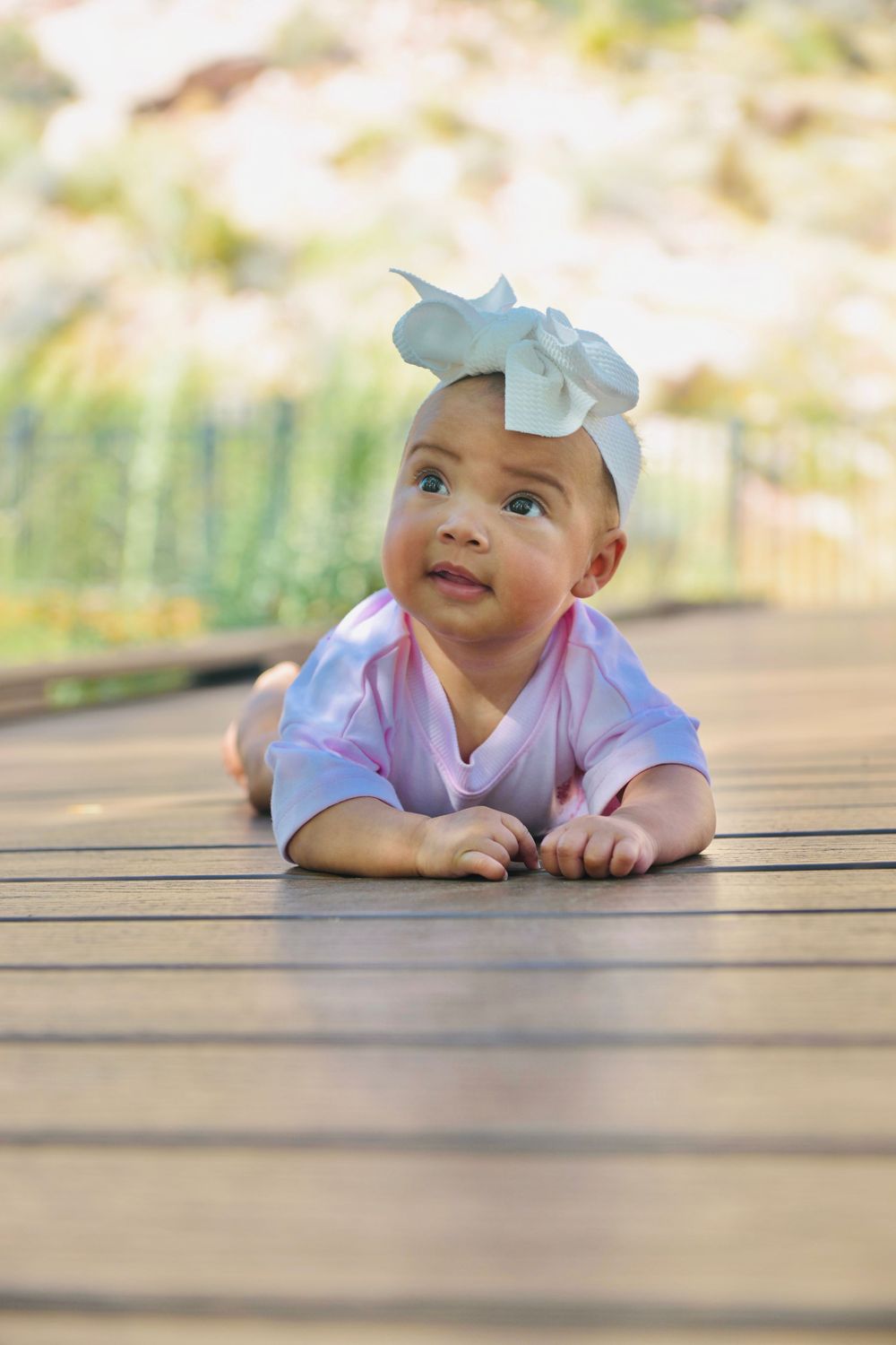 Adorable baby girl in pink onesie and white bow headband doing tummy time on a wooden deck outdoors.
