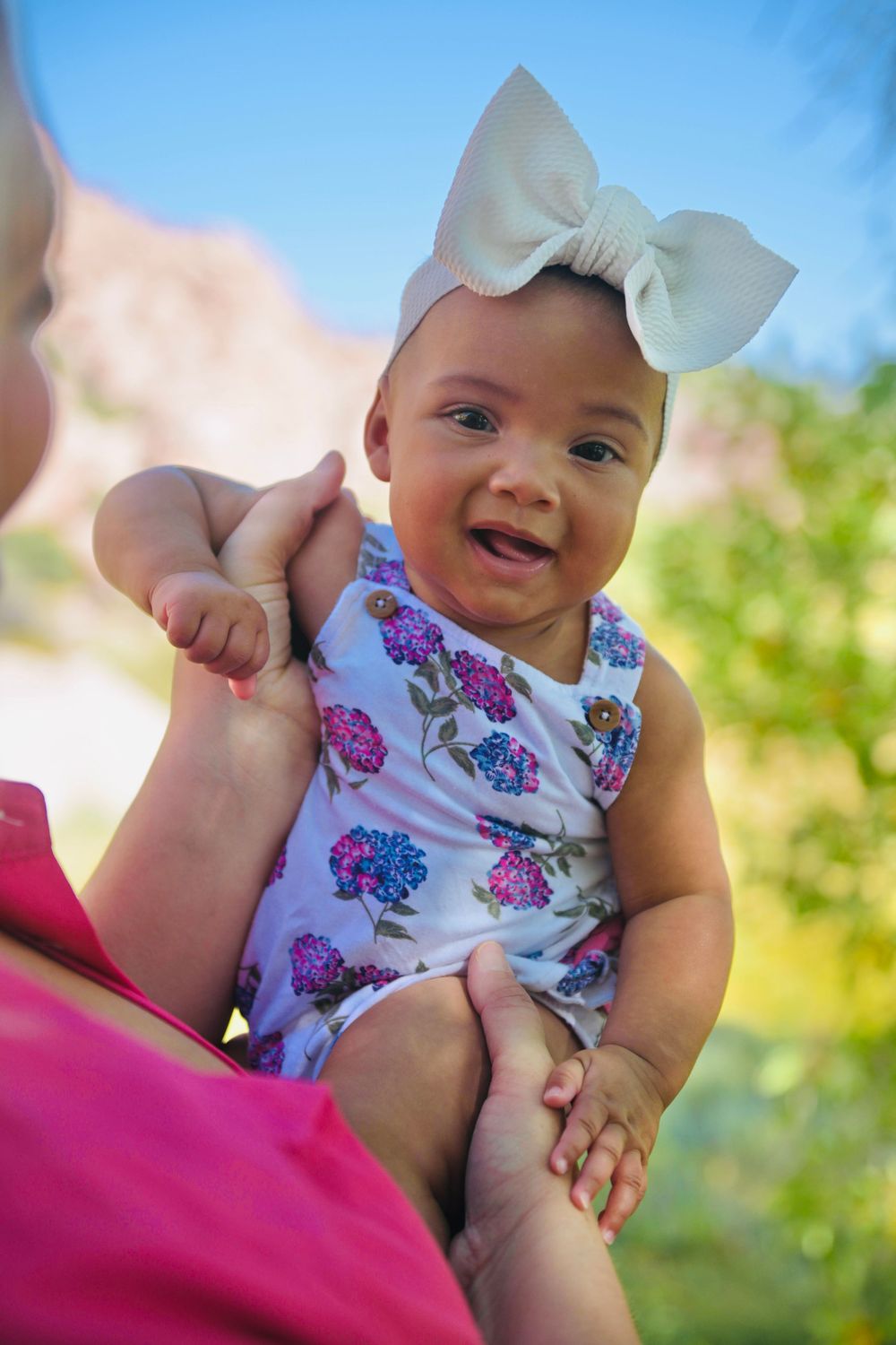 Smiling baby girl in floral romper and white bow headband being held outdoors on a sunny day.