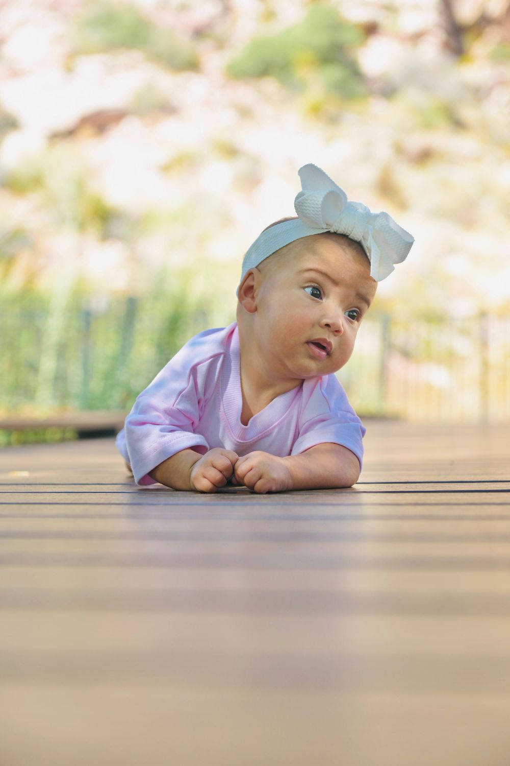 Adorable baby girl in pink outfit and blue bow headband lying on wooden deck, looking up curiously outdoors.