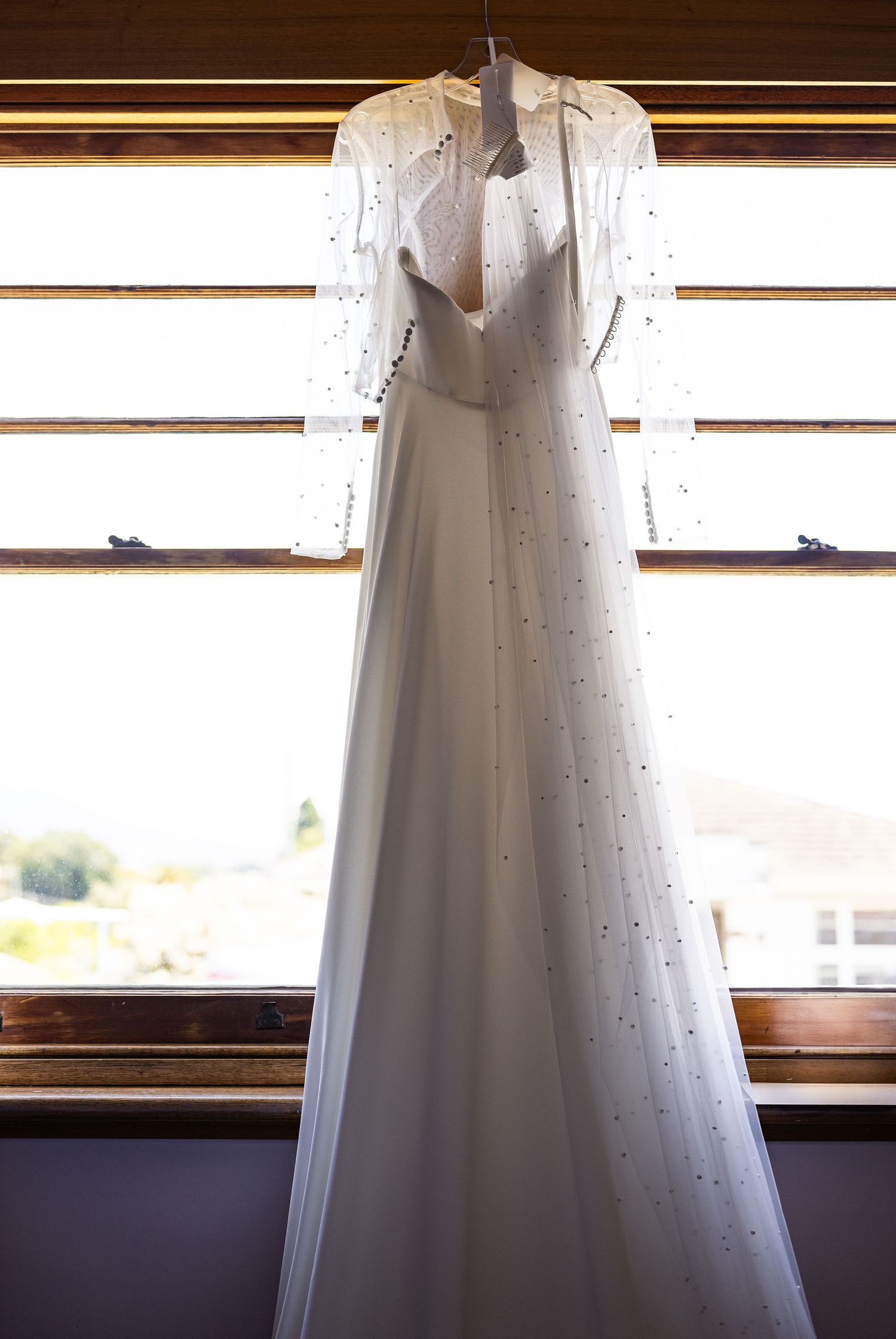 Elegant white wedding gown with pearl embellishments and sheer cape sleeves hanging by a sunlit window.