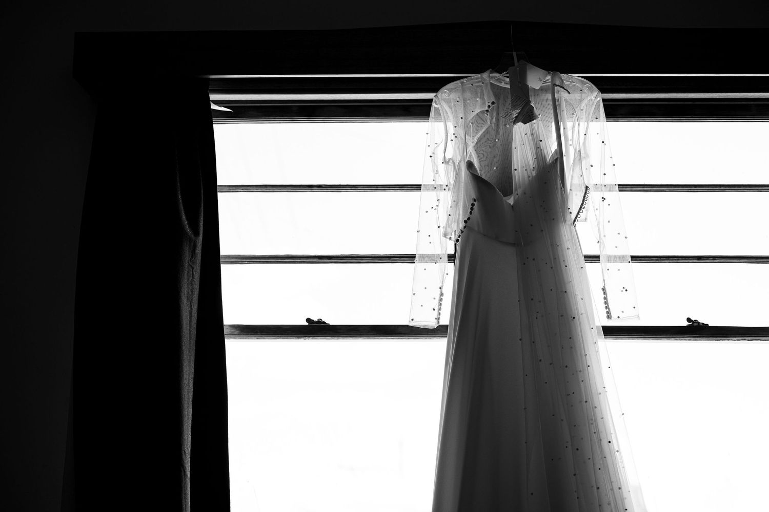Black and white photo of an elegant wedding dress hanging in front of a window, showcasing lace sleeves and flowing skirt.