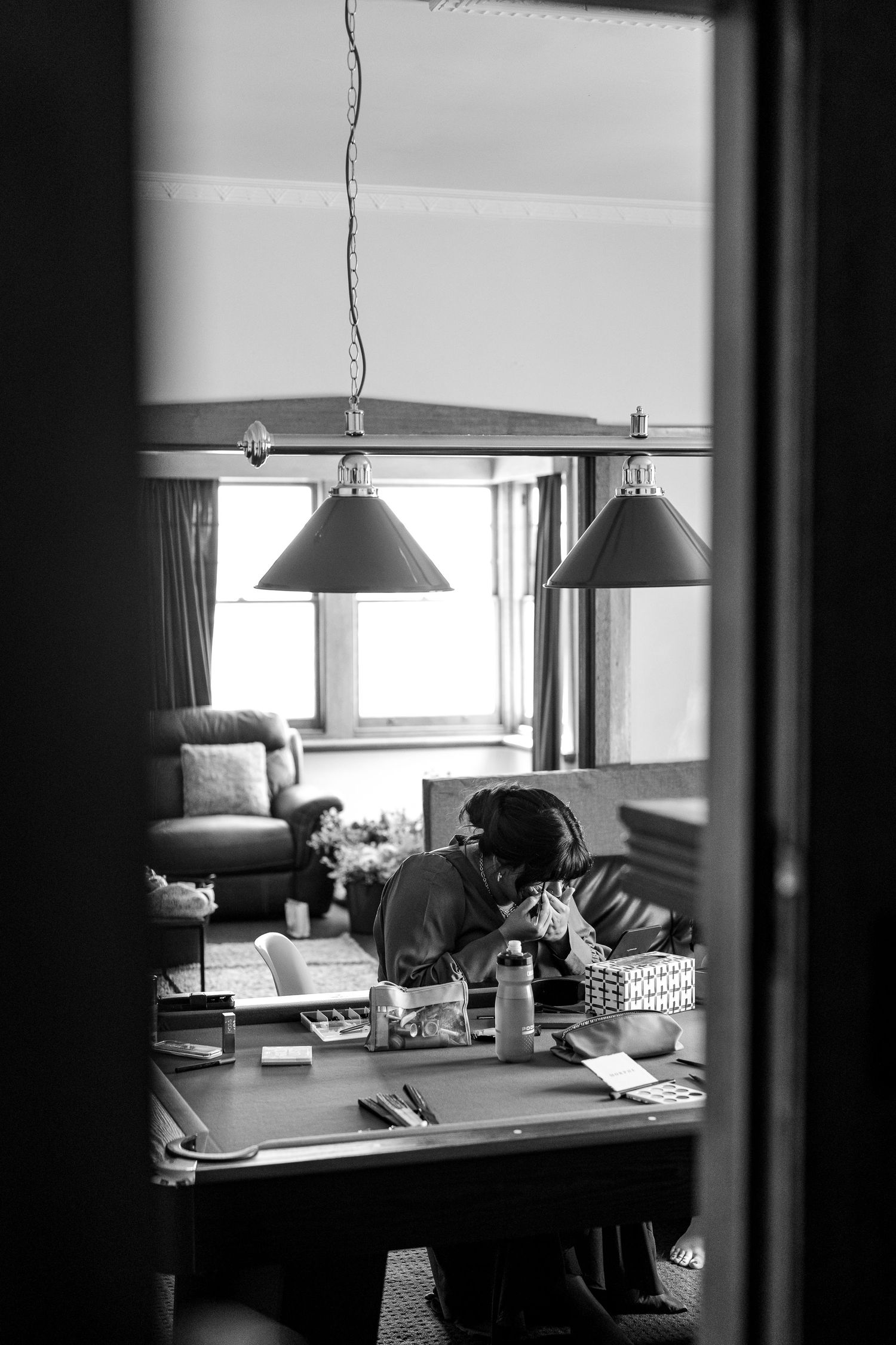 Black and white photo of a person sitting at a desk under pendant lamps, viewed through a doorway.