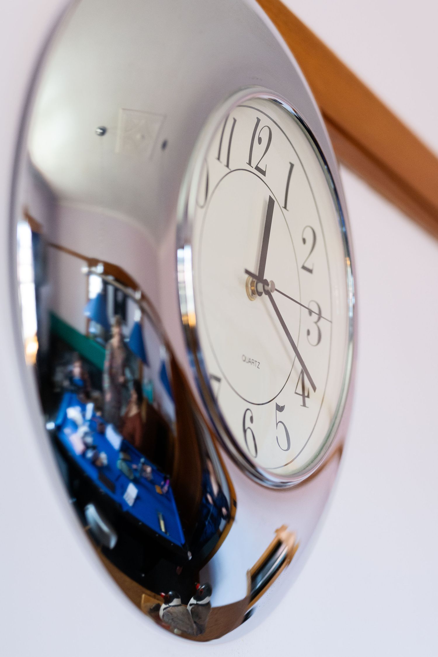 A silver oval wall clock with white face showing 3:20, reflecting a colorful room scene in its polished chrome frame.
