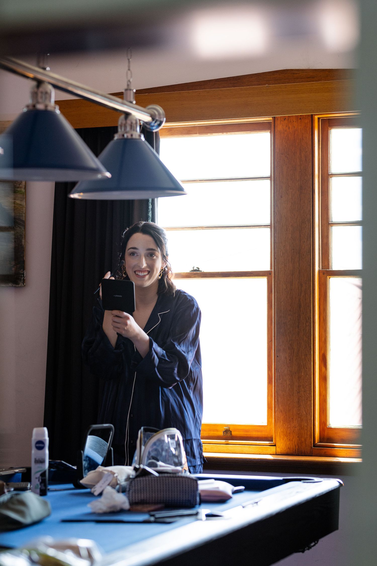 Woman in dark pajamas smiling while holding phone near window with pendant lights and vanity table.