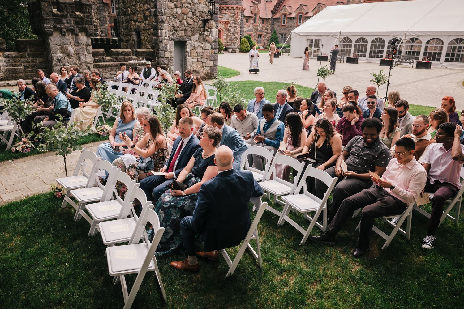 Wide view of seated wedding guests on the lawn at Searles Castle, with the officiant approaching in the background.