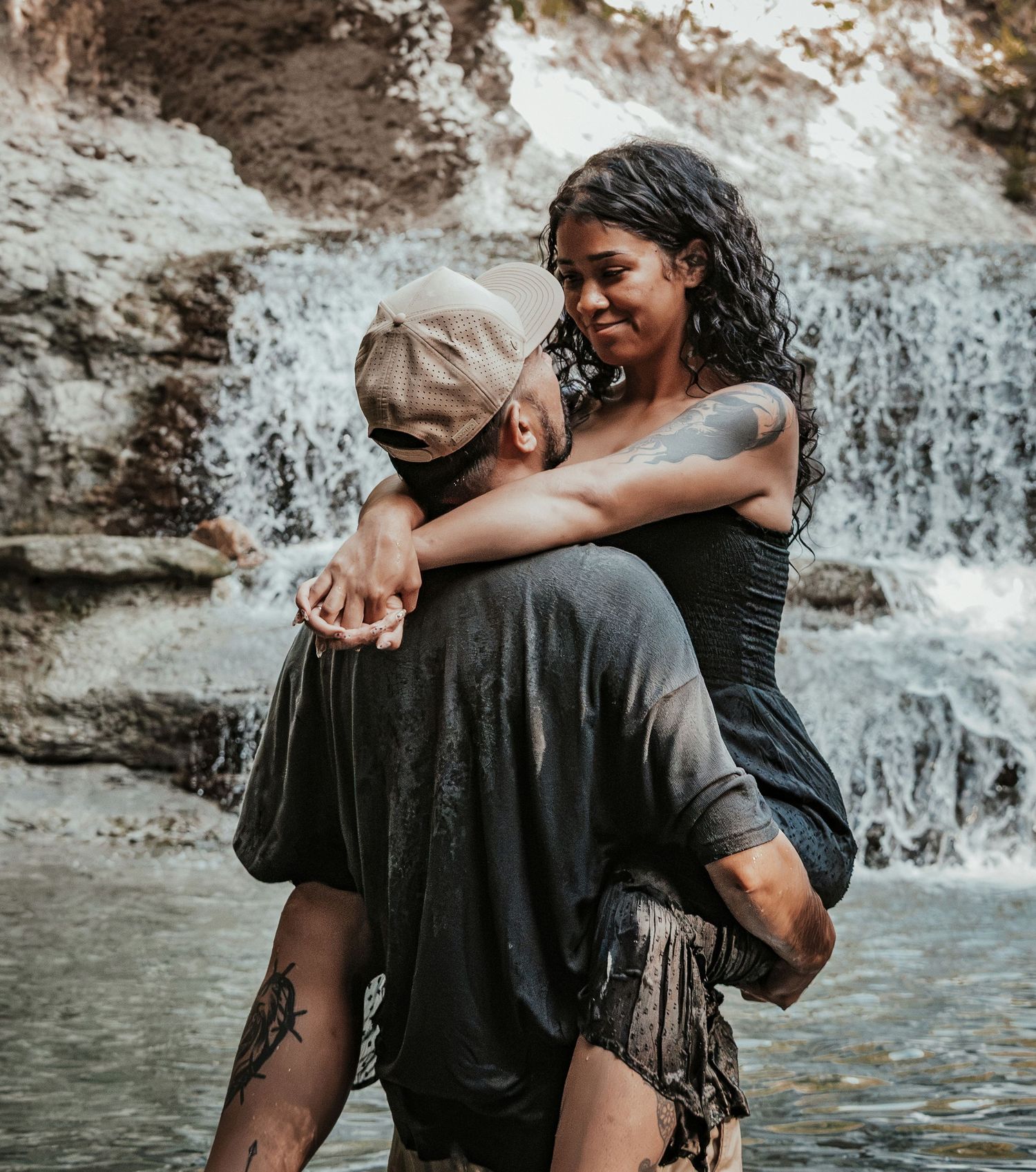 A couple embraces and smiles near a forest lake, the man in a white cap and gray shirt, woman in a black top.
