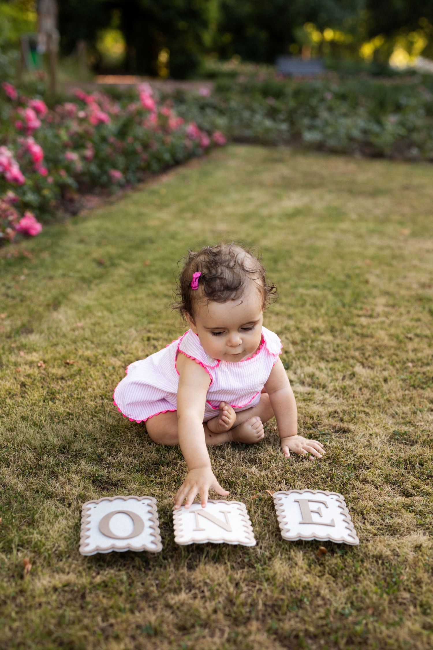 Baby girl in pink outfit sitting on grass reaching for letter tiles spelling ONE, celebrating her first birthday outdoors.