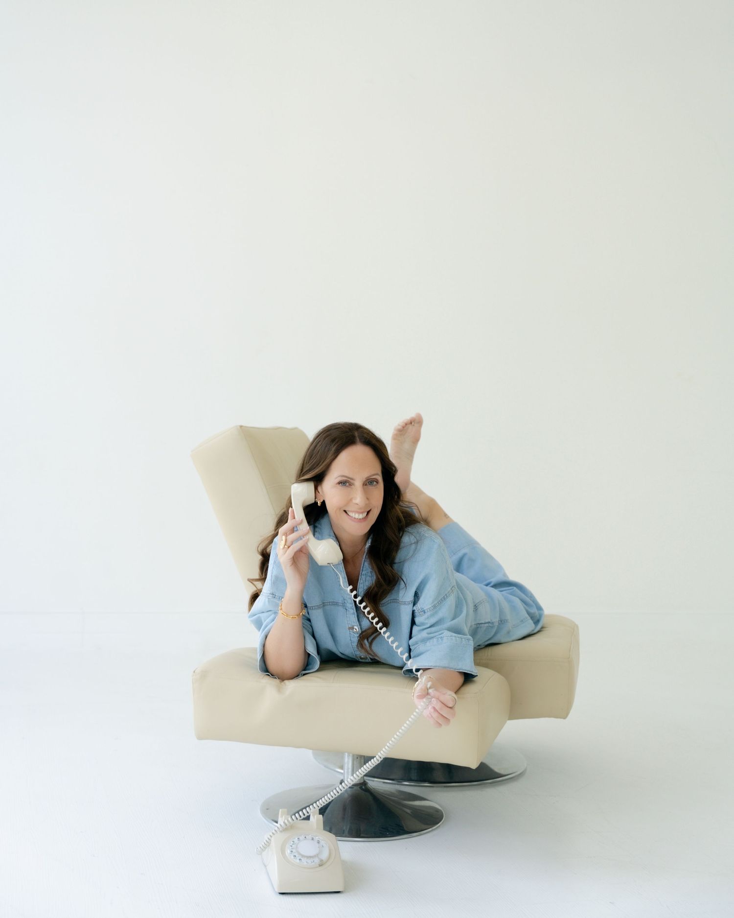 Woman in white sleeveless shirt and jeans holding a phone, smiling against a white draped fabric background.