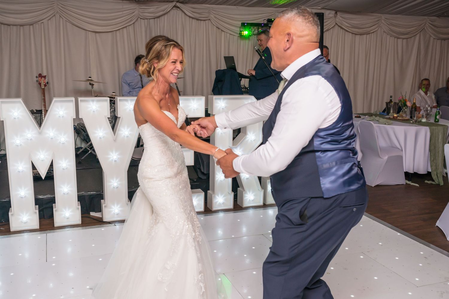 Bride and groom share their first dance on a white LED dance floor at a wedding reception with illuminated letter decorations.