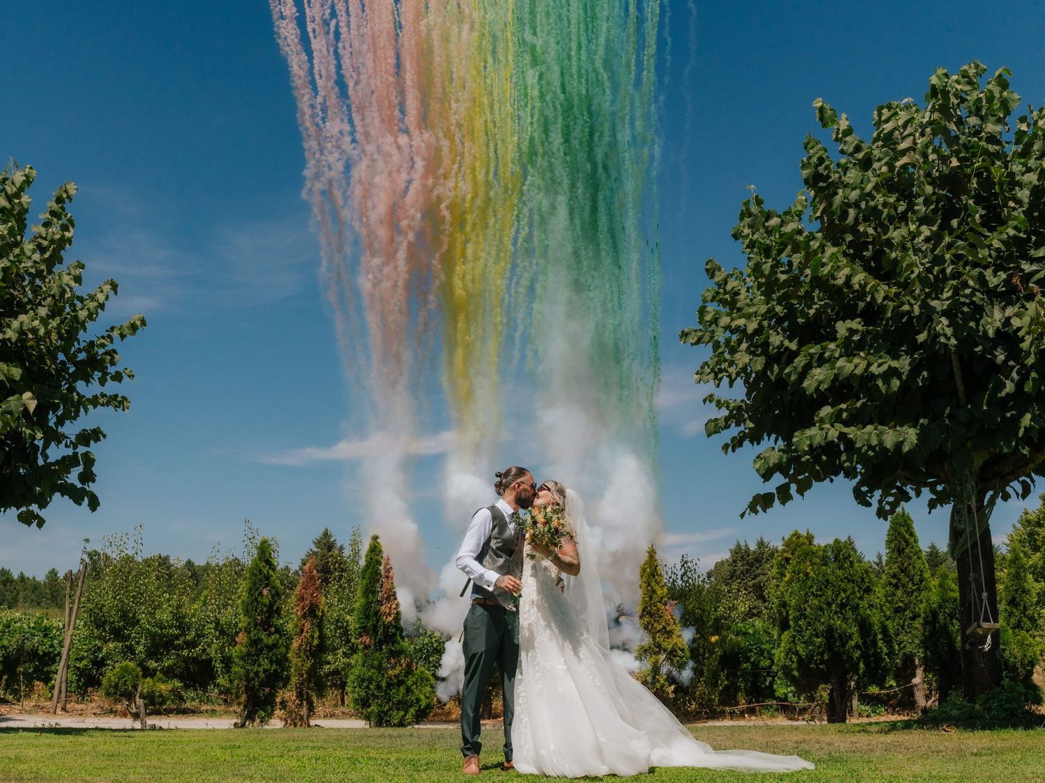 Bride and groom kissing outdoors with colorful rainbow smoke bombs exploding above them against a blue sky.