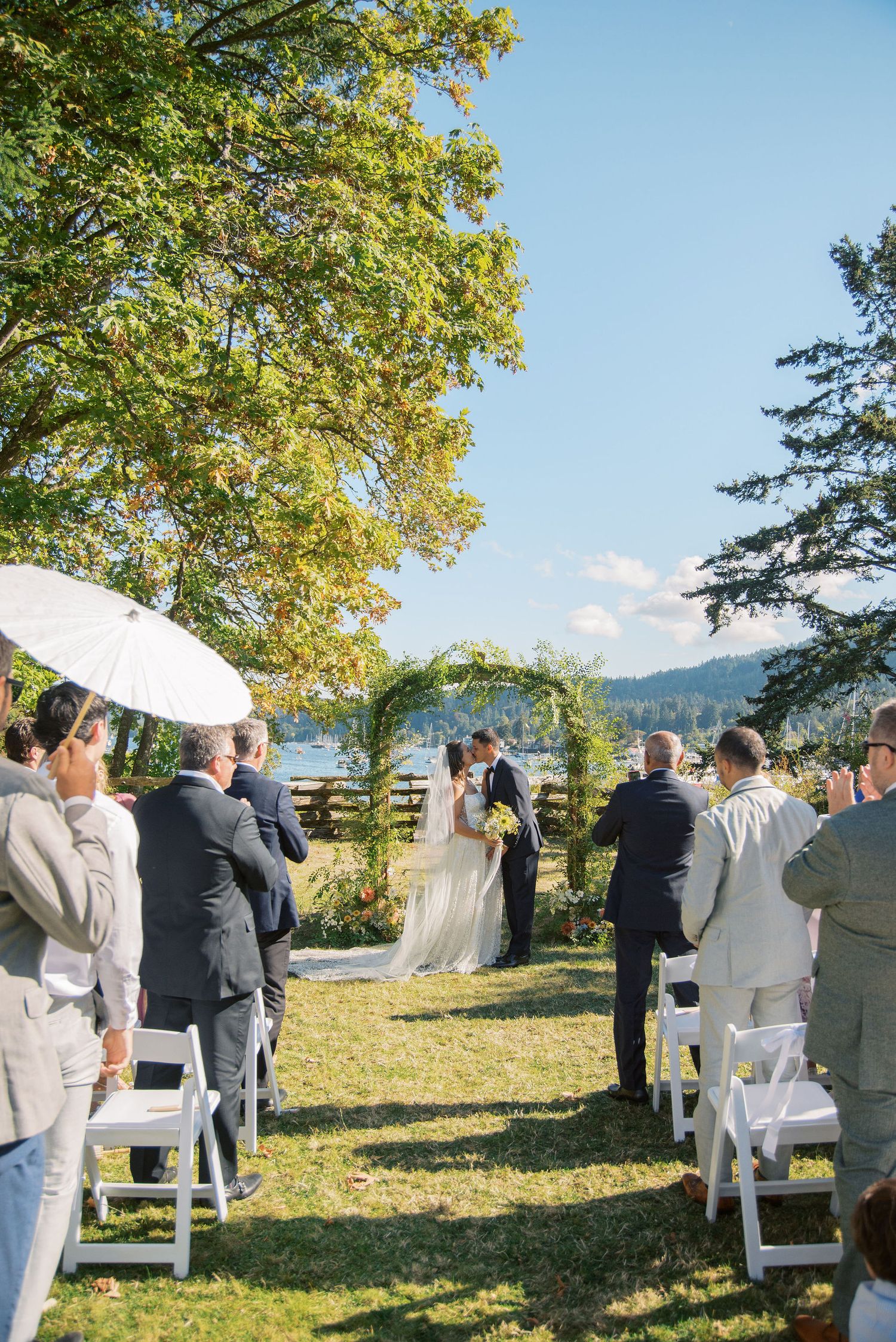 Outdoor wedding ceremony on green lawn with guests seated in white chairs, couple kissing at floral arch near lakeside.