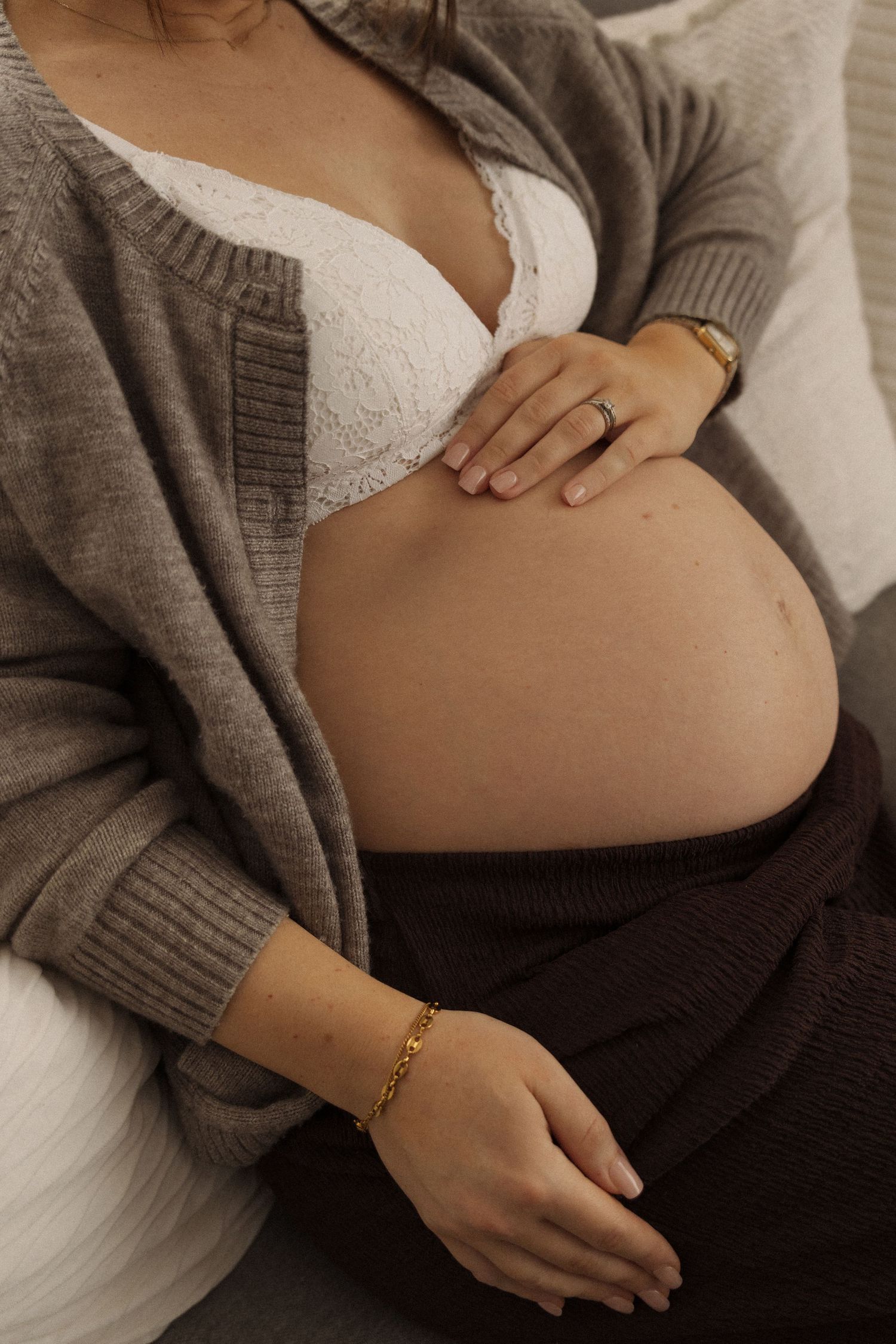 Pregnant woman in white lace bra and brown cardigan cradling baby bump, wearing gold bracelet and ring.