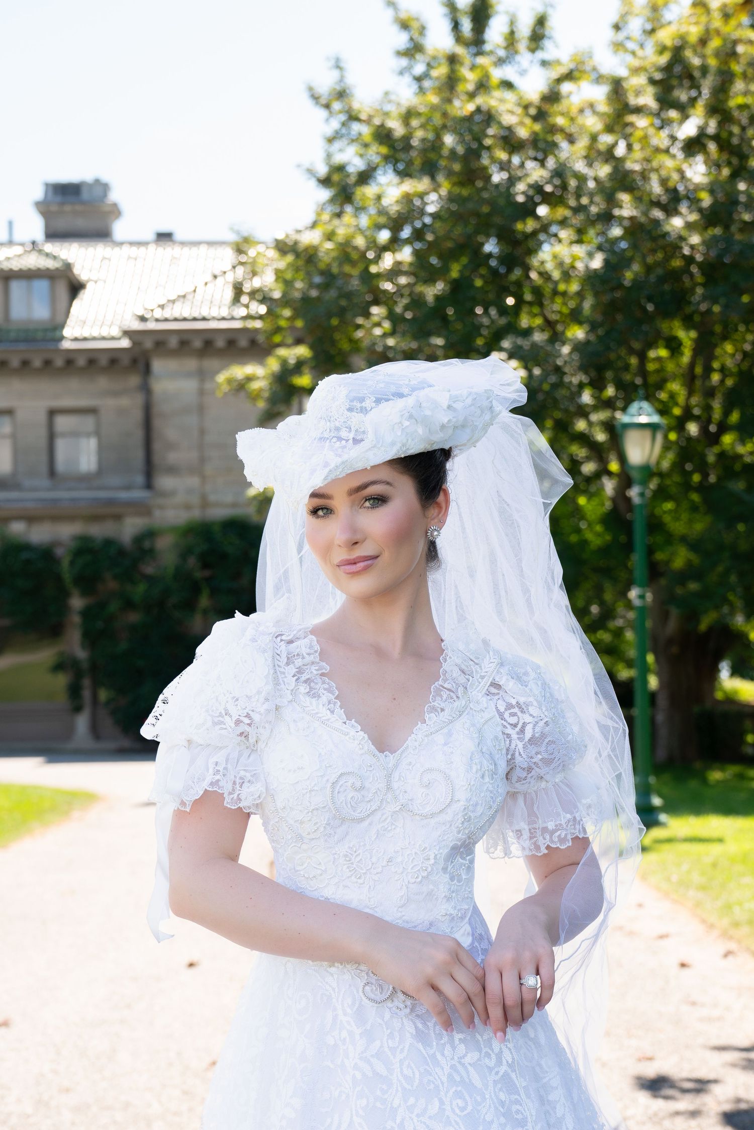 Beautiful bride in vintage white lace gown and matching hat with veil poses outdoors near a historic estate.