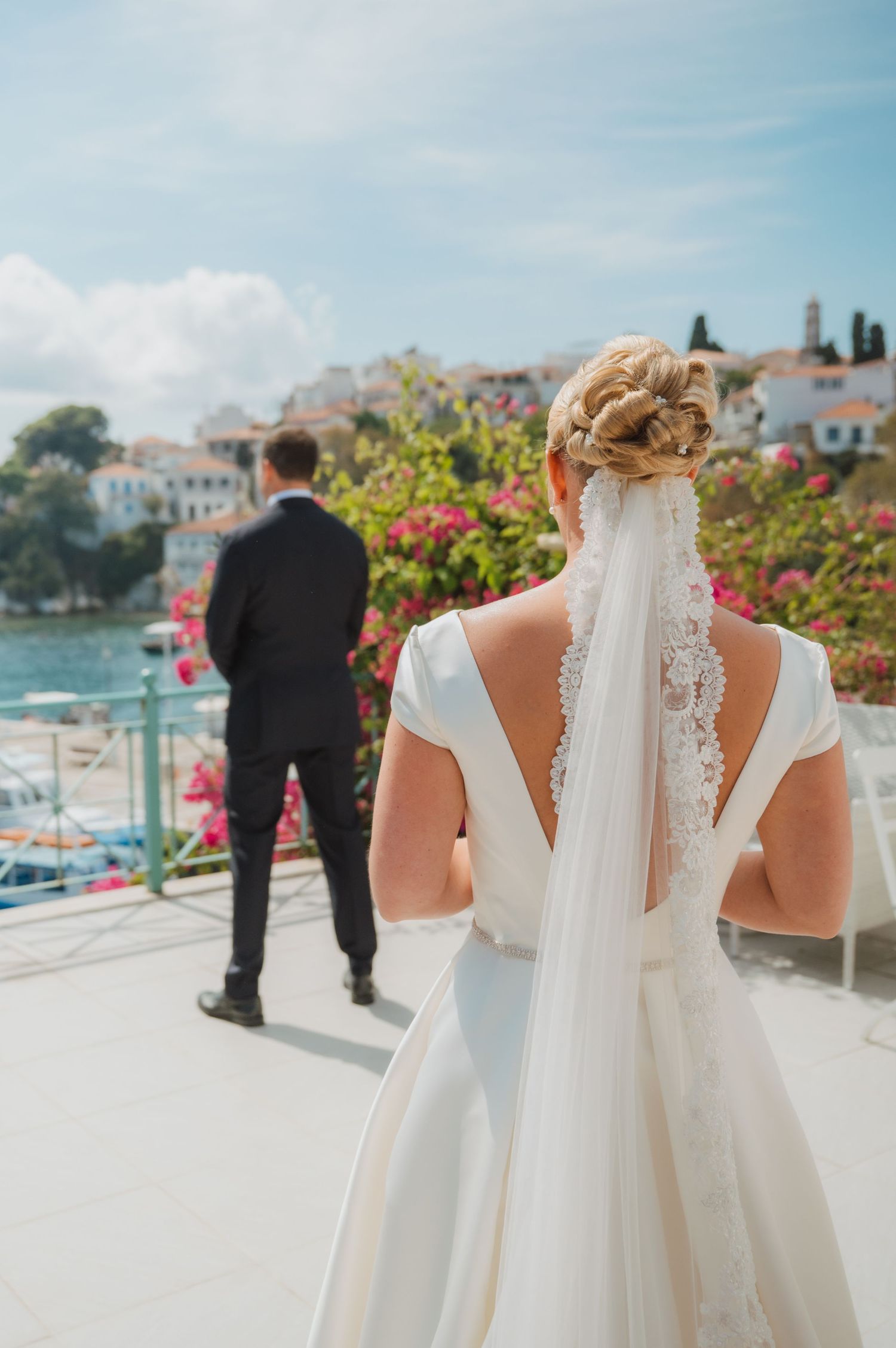 Bride in white gown with lace veil stands before groom overlooking a scenic Mediterranean coastal town.