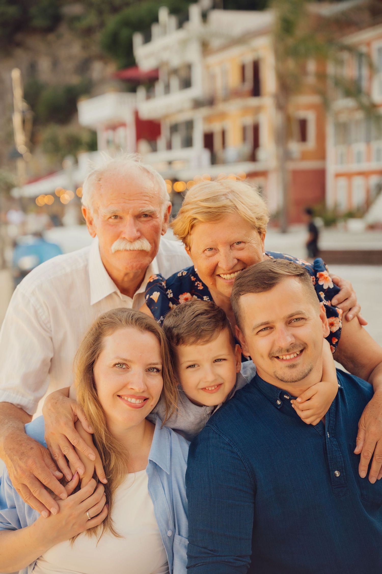 Multi-generational family of five smiling together outdoors near colorful Mediterranean buildings at sunset.
