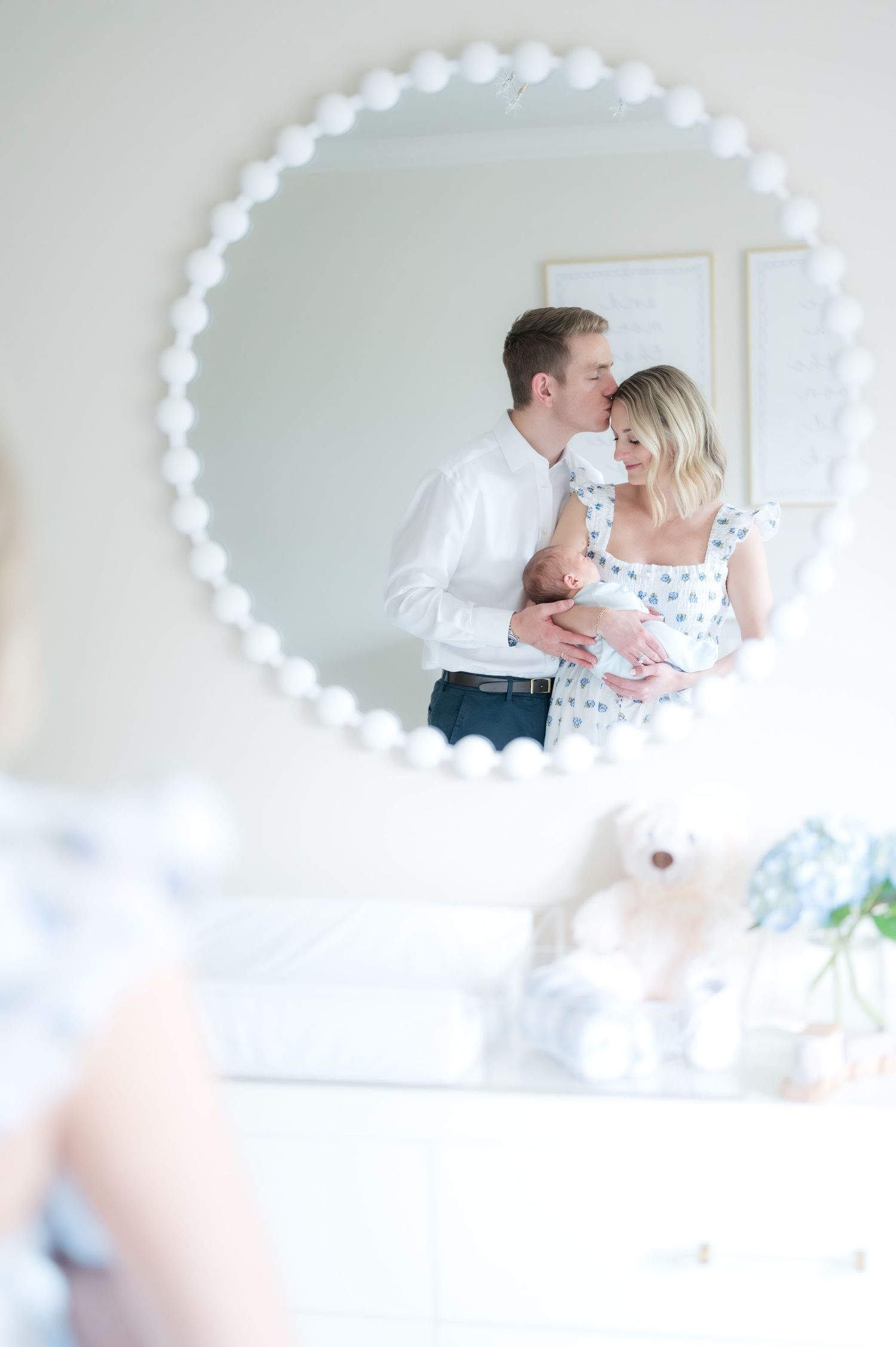 A father kissing a mother holding a newborn baby with the image reflection from a mirror in the baby's nursery.