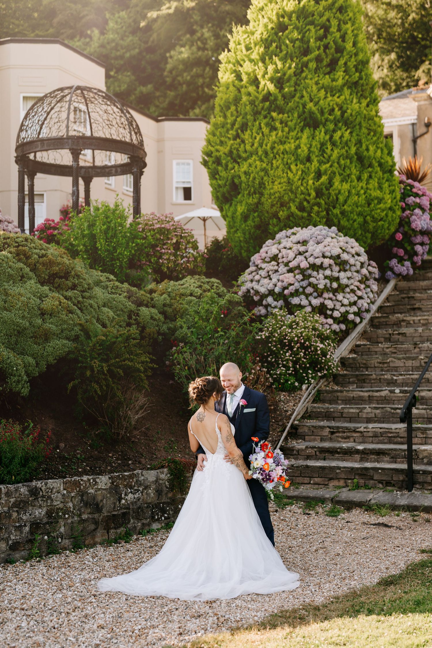 A romantic wedding couple shares a tender moment in front of a rustic white building with arched black doors.
